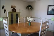 Round wooden dining table with four white spindle-back chairs and a vase of flowers in a simple communal room with cabinets and wall decorations.