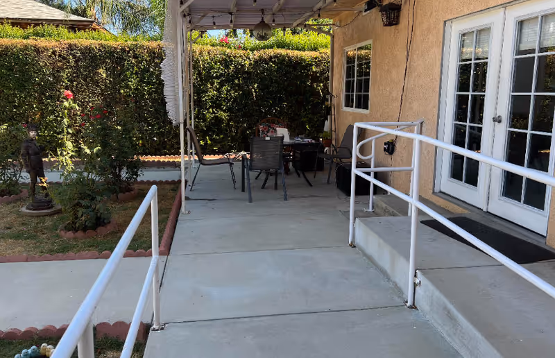 Outdoor patio area with a concrete ramp and steps leading to a building entrance with double glass doors. The patio has a canopy overhead, a table with chairs, and is surrounded by greenery including a hedge and some plants. There is a small statue visible in the garden area.