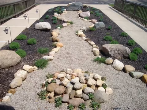 A landscaped outdoor garden area with a gravel pathway bordered by rocks and small green plants. The pathway is flanked by two concrete walkways with black lamp posts along the edges. Large rocks are placed decoratively within the garden beds covered with dark mulch.
