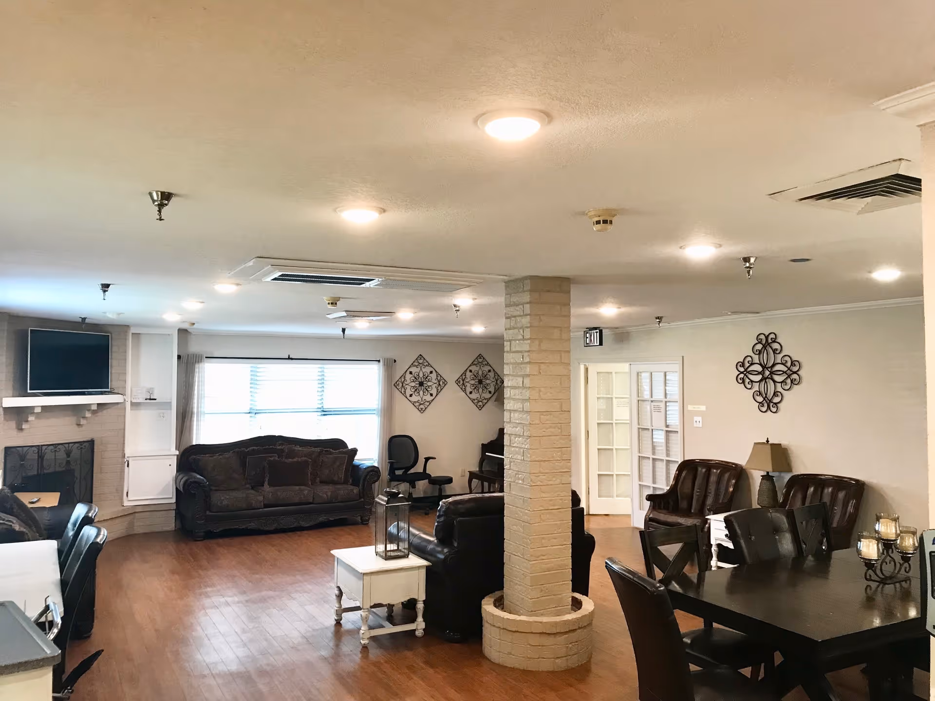 A spacious living room area in Oak Grove Nursing Home featuring multiple dark leather sofas and chairs arranged around a central brick column. The room has wooden flooring, a fireplace with a mounted TV above it, a window with curtains, decorative wall art, and a dining table with chairs. The ceiling has multiple recessed lights and air vents.