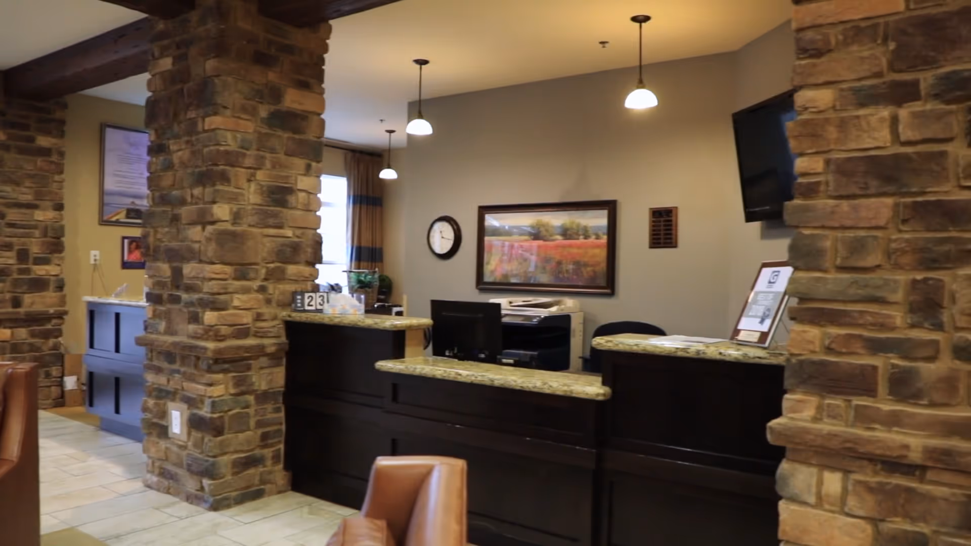 Reception area with a dark wood desk and granite countertop, stone pillars, a wall clock, a landscape painting, a mounted TV, and a few chairs in the foreground.