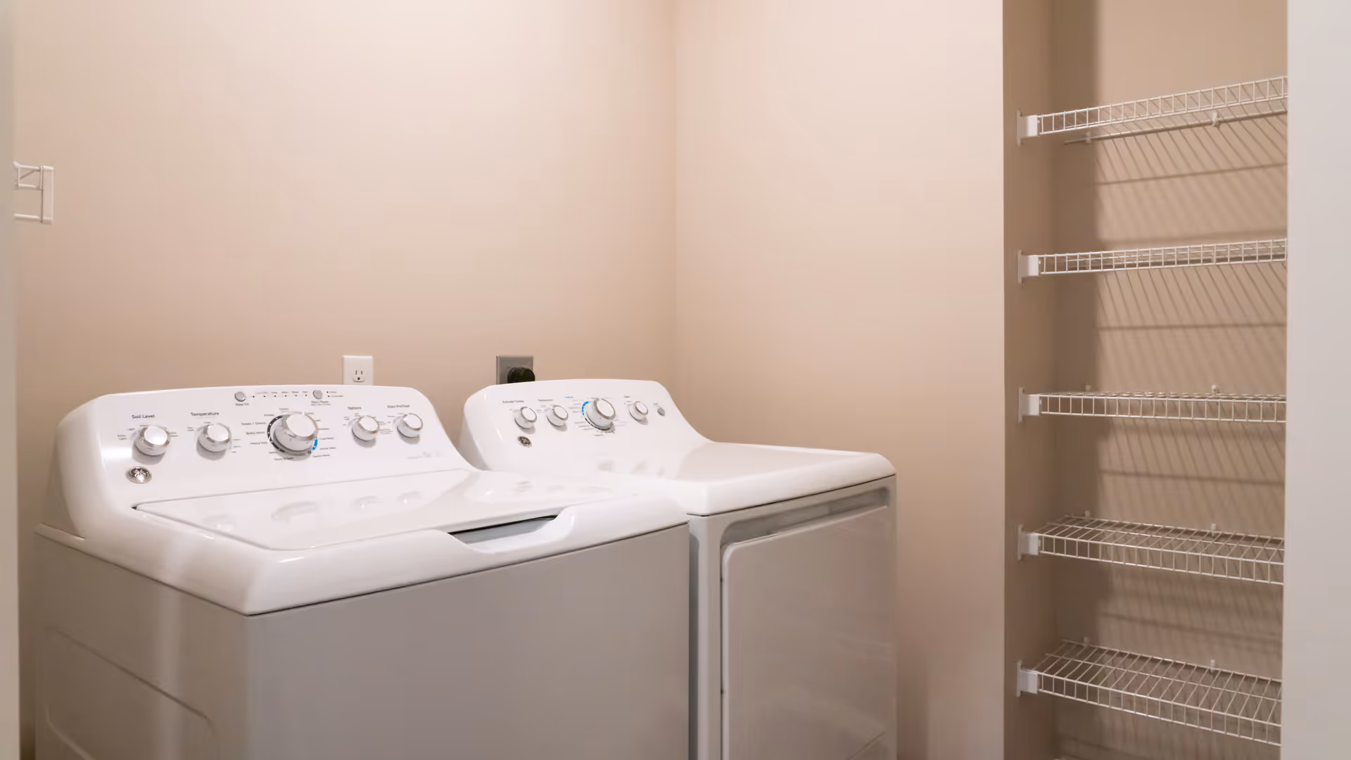A laundry room with a white top-loading washing machine and matching dryer beside wall-mounted wire shelving.