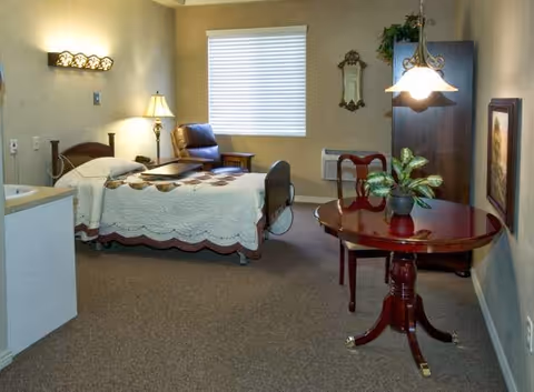 A cozy senior living room with a single bed covered in a white quilt, a wooden side table with a lamp, a brown leather armchair, a round wooden table with a plant on it, a hanging light fixture, a window with blinds, and a wall-mounted mirror and framed picture.