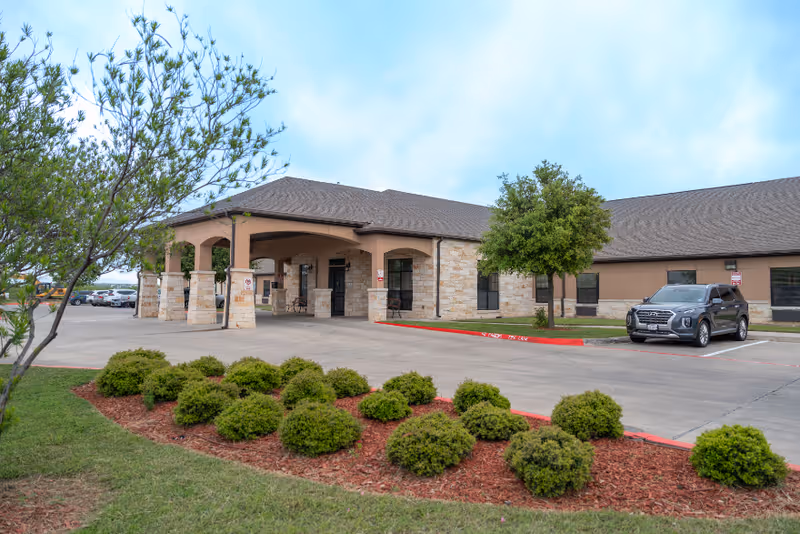 Exterior view of Laredo Nursing and Rehabilitation Center showing a single-story building with stone and beige walls, a covered entrance supported by stone pillars, a parking area with a parked SUV, and landscaped bushes and trees under a cloudy sky.