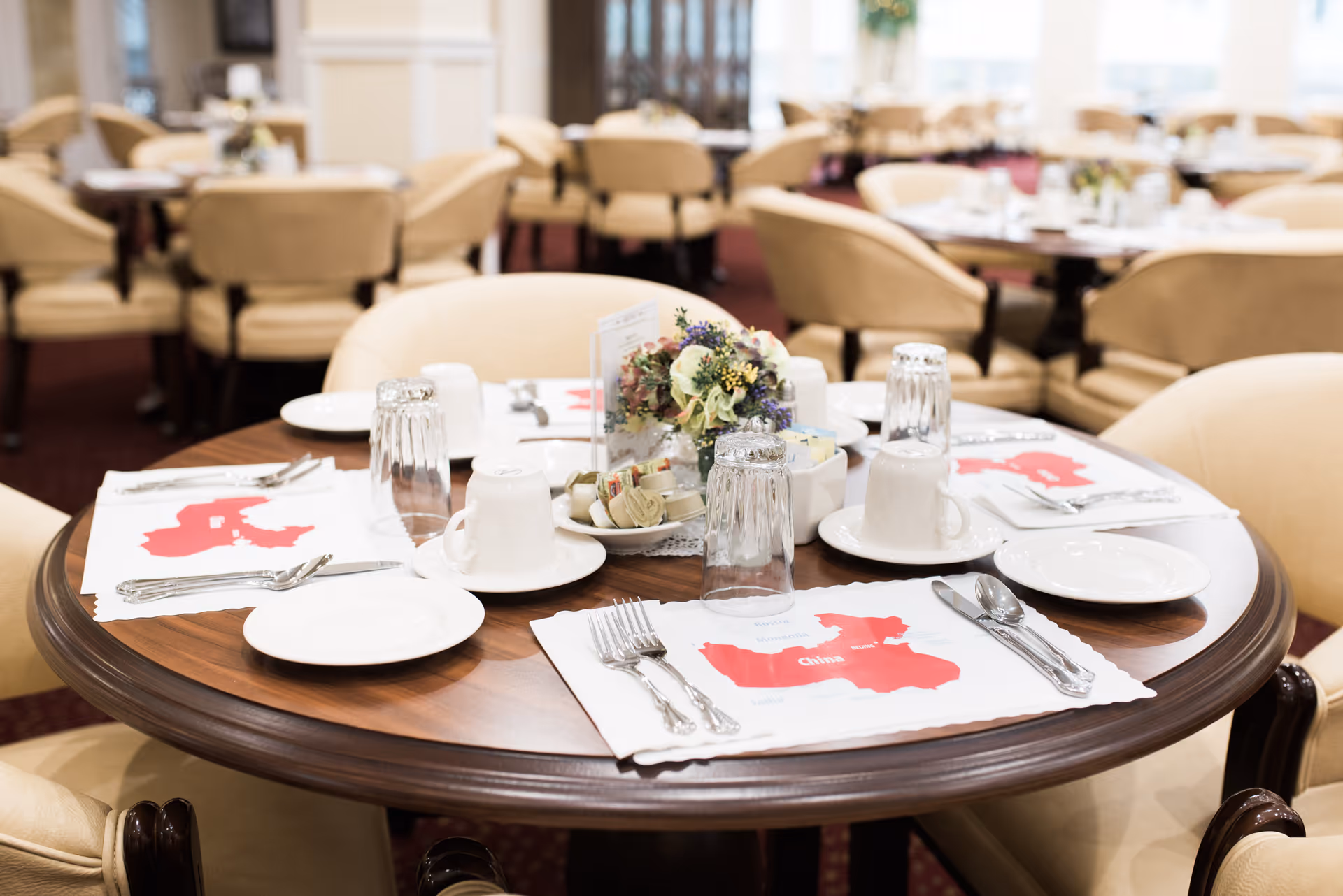 A round wooden dining table set for four with white plates, cups, silverware, and glasses. Each place setting has a placemat featuring a red map labeled 'China'. A small floral centerpiece and condiments are in the middle of the table. The background shows more tables and beige upholstered chairs in a softly lit dining room.