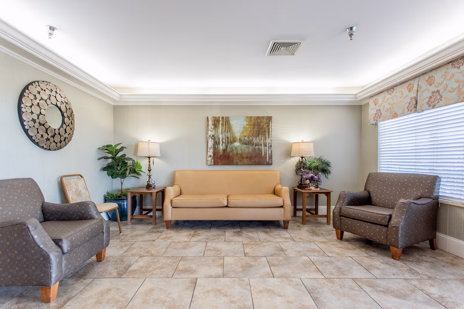 A bright and clean living room area in a nursing and rehab facility featuring a beige sofa centered against the wall, flanked by two wooden side tables with lamps and plants. Two patterned armchairs are placed on either side of the sofa, with a large window covered by blinds and floral valance on the right. The floor is tiled, and the walls are light-colored with a decorative round mirror and a framed painting above the sofa.