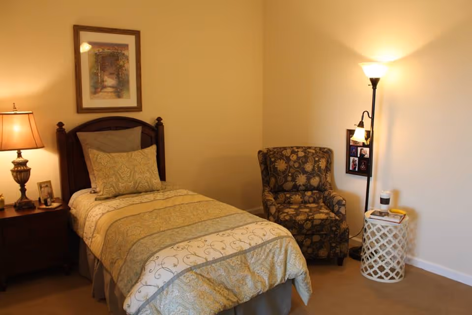 A cozy bedroom with a single bed featuring patterned bedding and pillows. To the left of the bed is a wooden nightstand with a lamp and framed photos. On the right side of the room, there is a floral upholstered armchair next to a white lattice side table holding a cup and a banana. A tall floor lamp with two lights stands behind the side table, and a framed picture hangs on the wall above the bed.