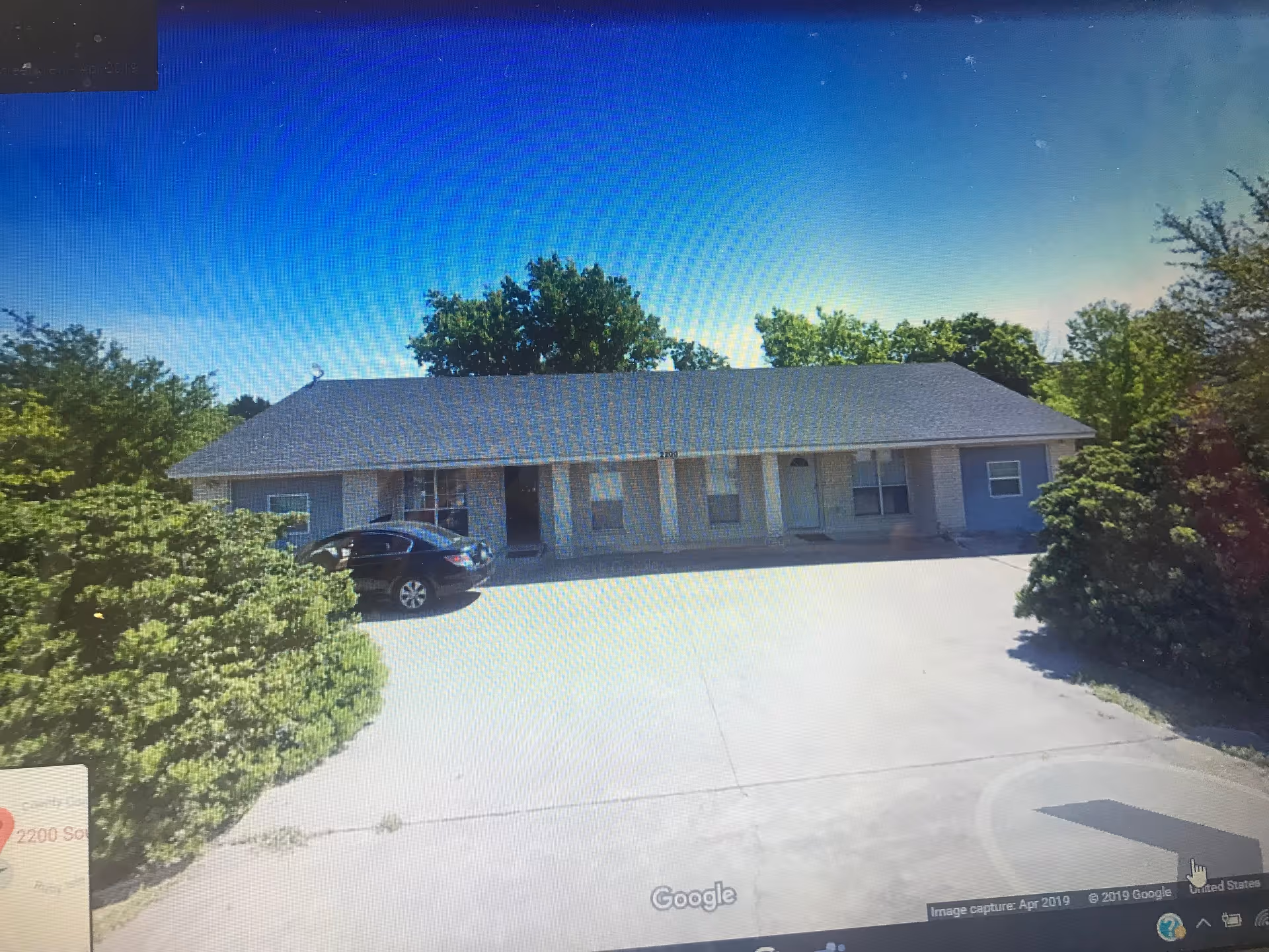 Single-story residential building with a gray roof and light-colored brick exterior, surrounded by green bushes and trees under a clear blue sky. A black car is parked in the driveway in front of the building.