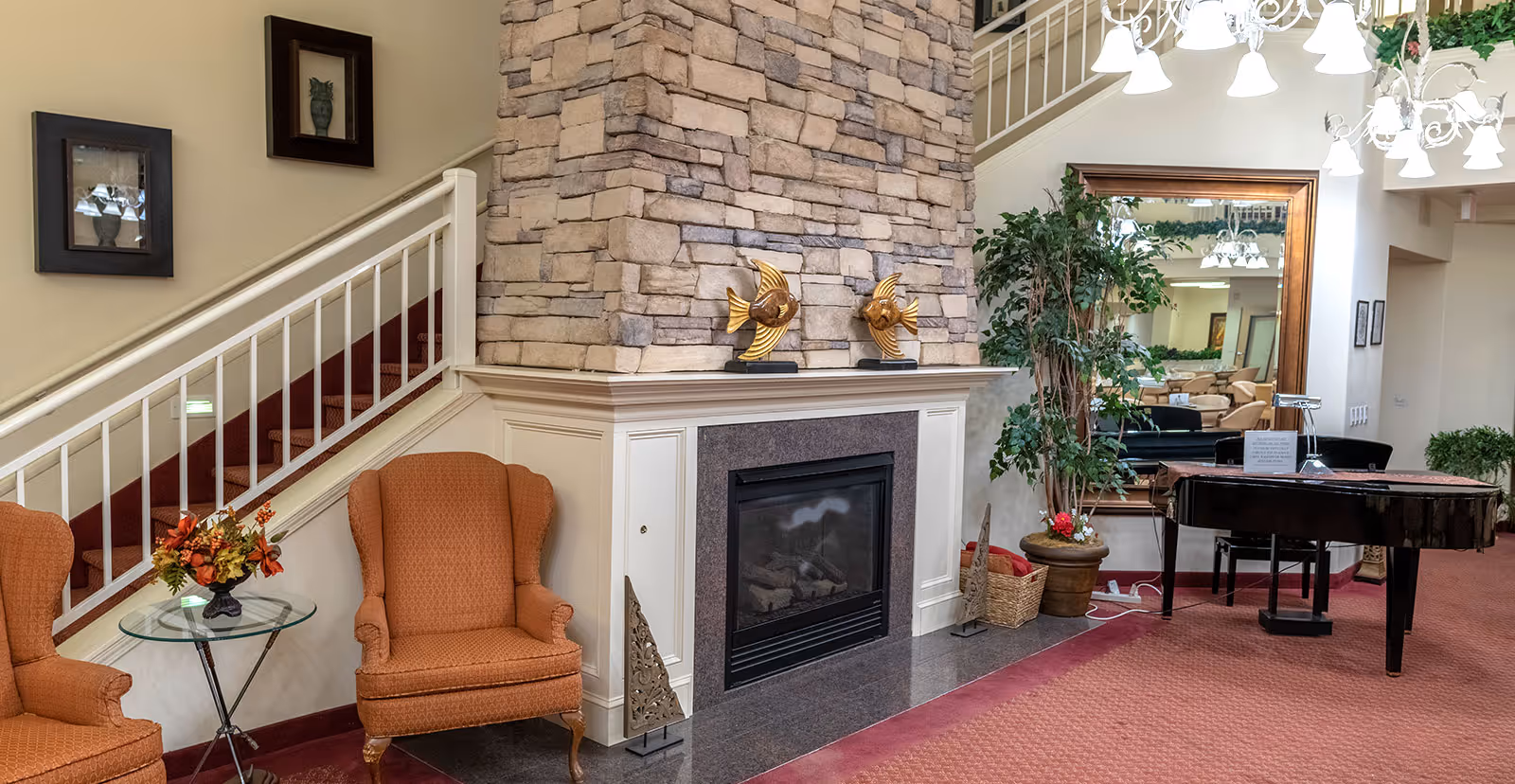 Cozy interior living area featuring a stone fireplace with two decorative fish sculptures on the mantel, two orange upholstered armchairs with a small glass side table holding a floral arrangement, a large mirror reflecting the room, a potted plant, and a black grand piano under a chandelier with multiple lights.
