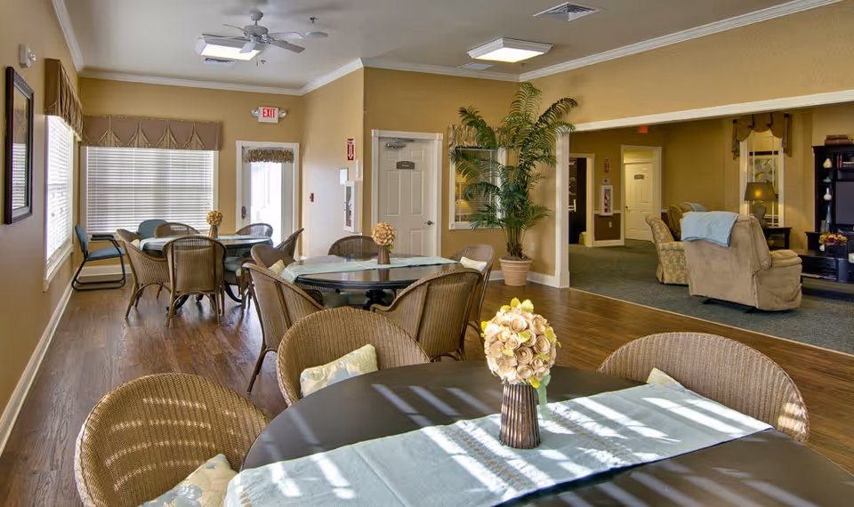 Communal dining room with round tables, wicker chairs, floral centerpieces, and a sitting area in the background.