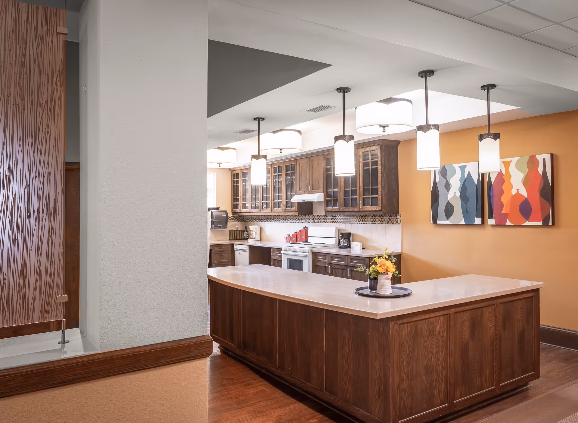 A spacious kitchen area with a large wooden island countertop in the center. The kitchen features dark wooden cabinets with glass doors, a white stove and dishwasher, and modern pendant lights hanging from the ceiling. The walls are painted a warm orange color, and two abstract art pieces are displayed on the wall. A small flower arrangement is placed on the island countertop.