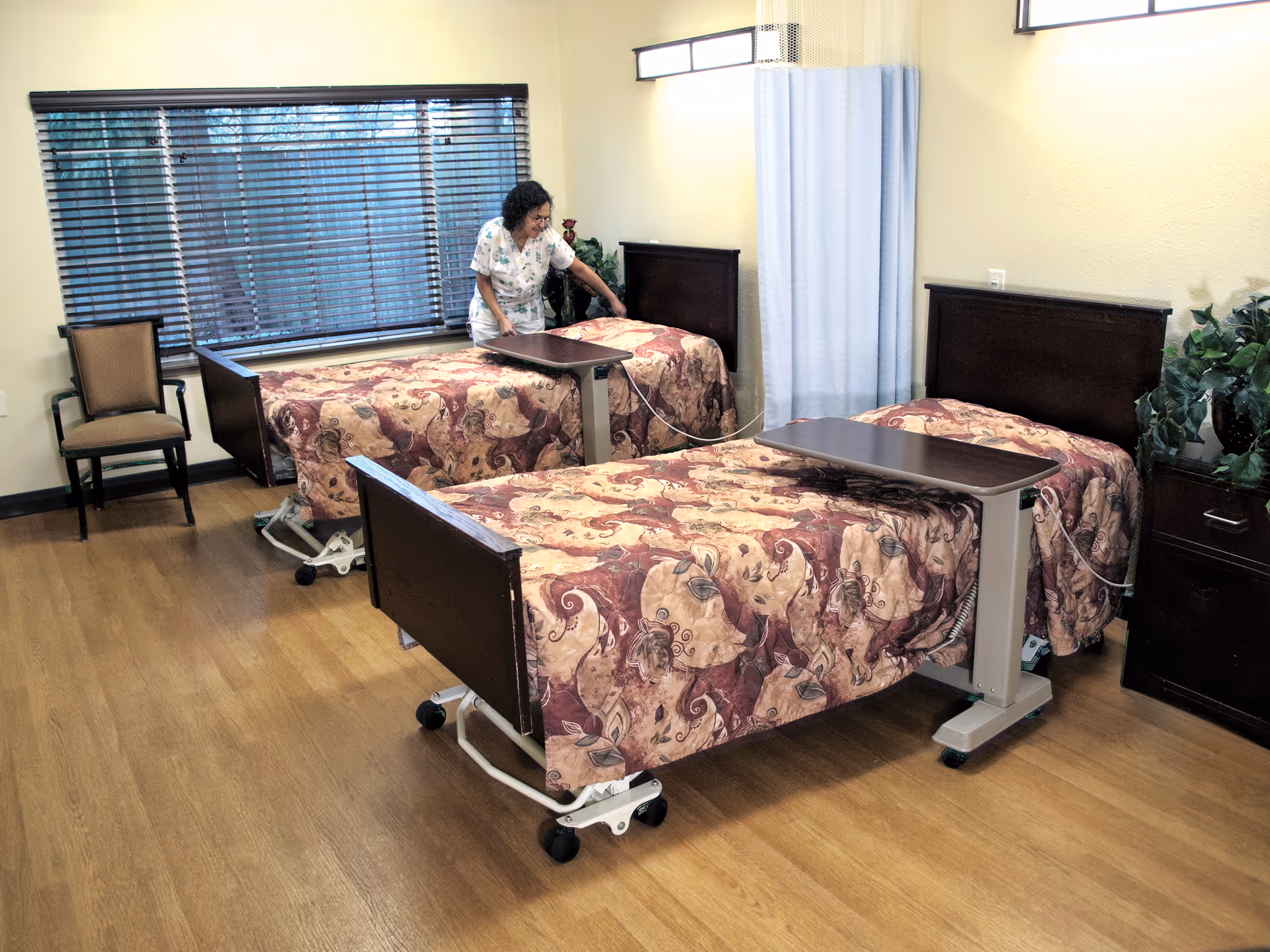A caregiver adjusts bedding in a shared patient bedroom with three hospital-style beds, overbed tables, wood floors, and a window with blinds.