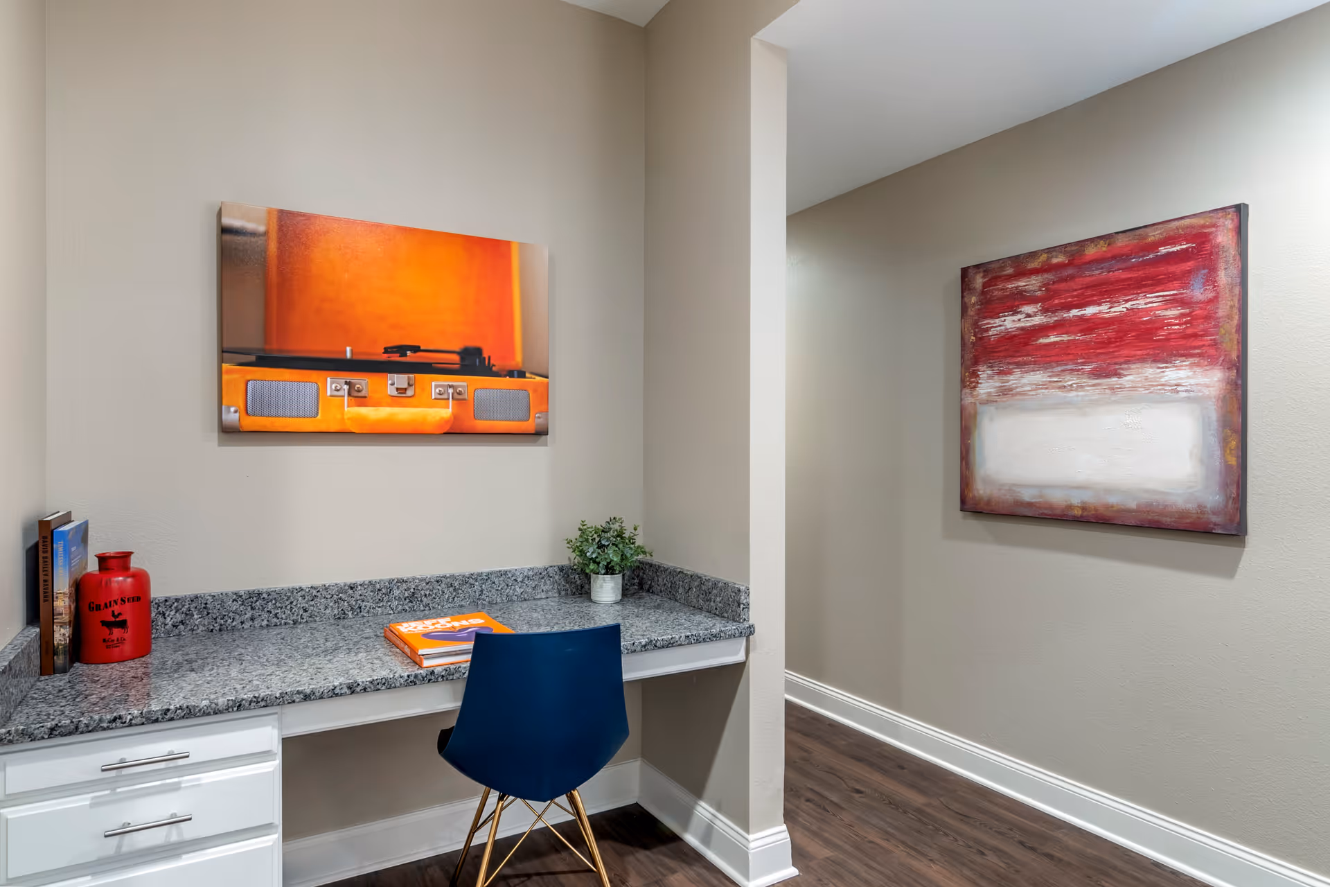 A small workspace with a granite countertop desk, a blue chair, and decorative items including books, a red vase, and a small plant. On the wall above the desk is a colorful painting of an orange record player. To the right, there is a hallway with a large abstract red and white painting on the wall. The floor is dark wood and the walls are painted light beige.