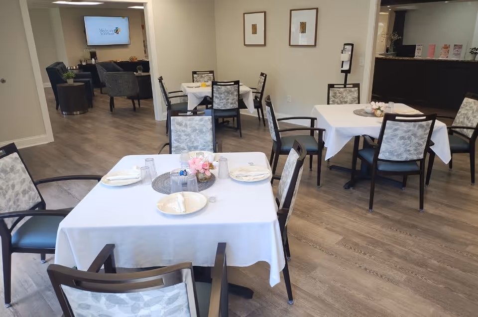 A dining room in a senior living facility with several tables covered in white tablecloths, each set with plates, napkins, and glasses. The chairs have patterned upholstery. In the background, there is a lounge area with armchairs and a TV mounted on the wall displaying the facility's logo. The floor is wood, and the walls are light-colored with framed artwork.