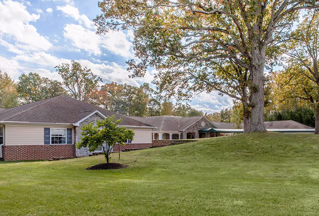 Single-story senior living building with brick and siding exterior, a large grassy front lawn and a mature tree under a partly cloudy sky.