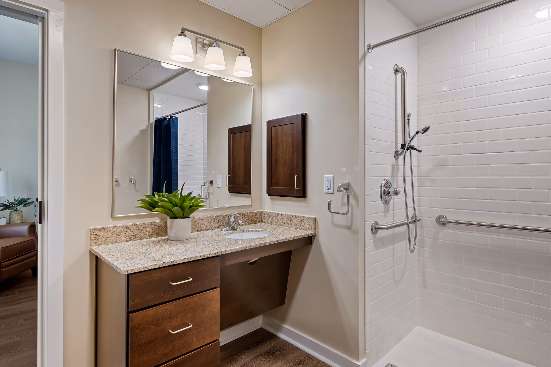 A modern bathroom with a granite countertop vanity featuring a sink, a potted green plant, and a large mirror above. There is a three-light fixture above the mirror. To the right is a walk-in shower with white subway tiles, a handheld showerhead, and multiple grab bars for accessibility. The bathroom has beige walls and wood flooring. A glimpse of an adjacent room with a brown chair and a lamp is visible through an open doorway on the left.