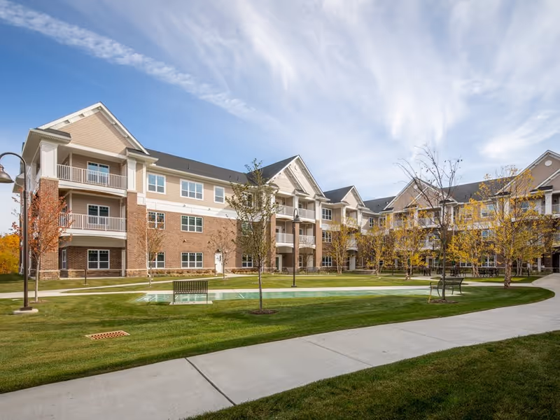 Exterior view of a multi-story senior living facility building with balconies, surrounded by a well-maintained lawn, young trees, benches, and a paved walkway under a partly cloudy sky.