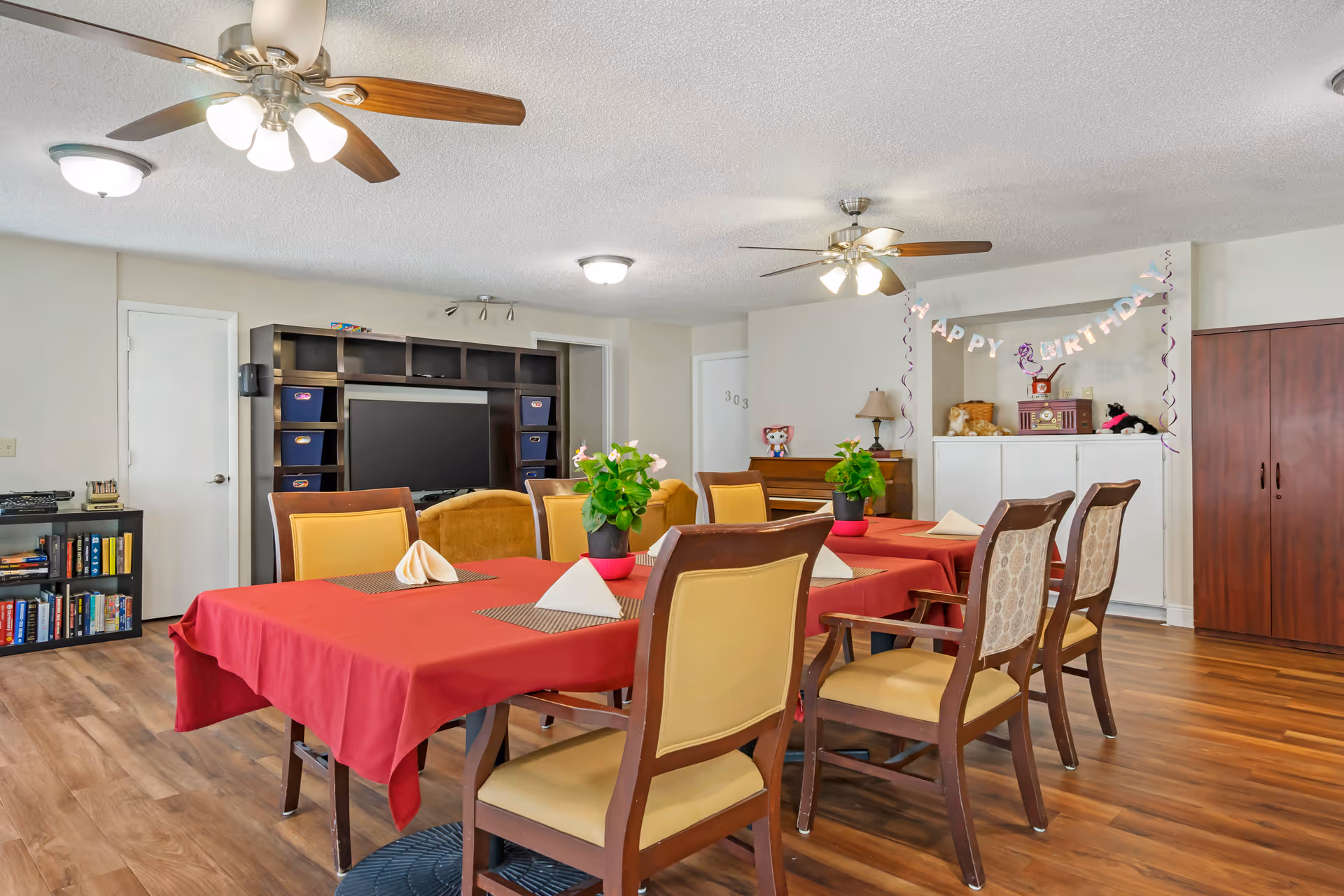 Communal dining/activity room with tables covered in red tablecloths, chairs, potted plants, a TV and a 'Happy Birthday' banner on the wall.