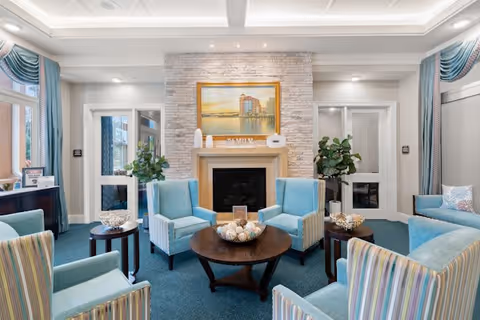 A cozy living room area with light blue and striped armchairs arranged around a dark wooden coffee table. The room features a stone fireplace with a painting above it, decorative items on the mantel, and large windows with blue curtains. There are green plants on either side of the fireplace and soft lighting from recessed ceiling lights.