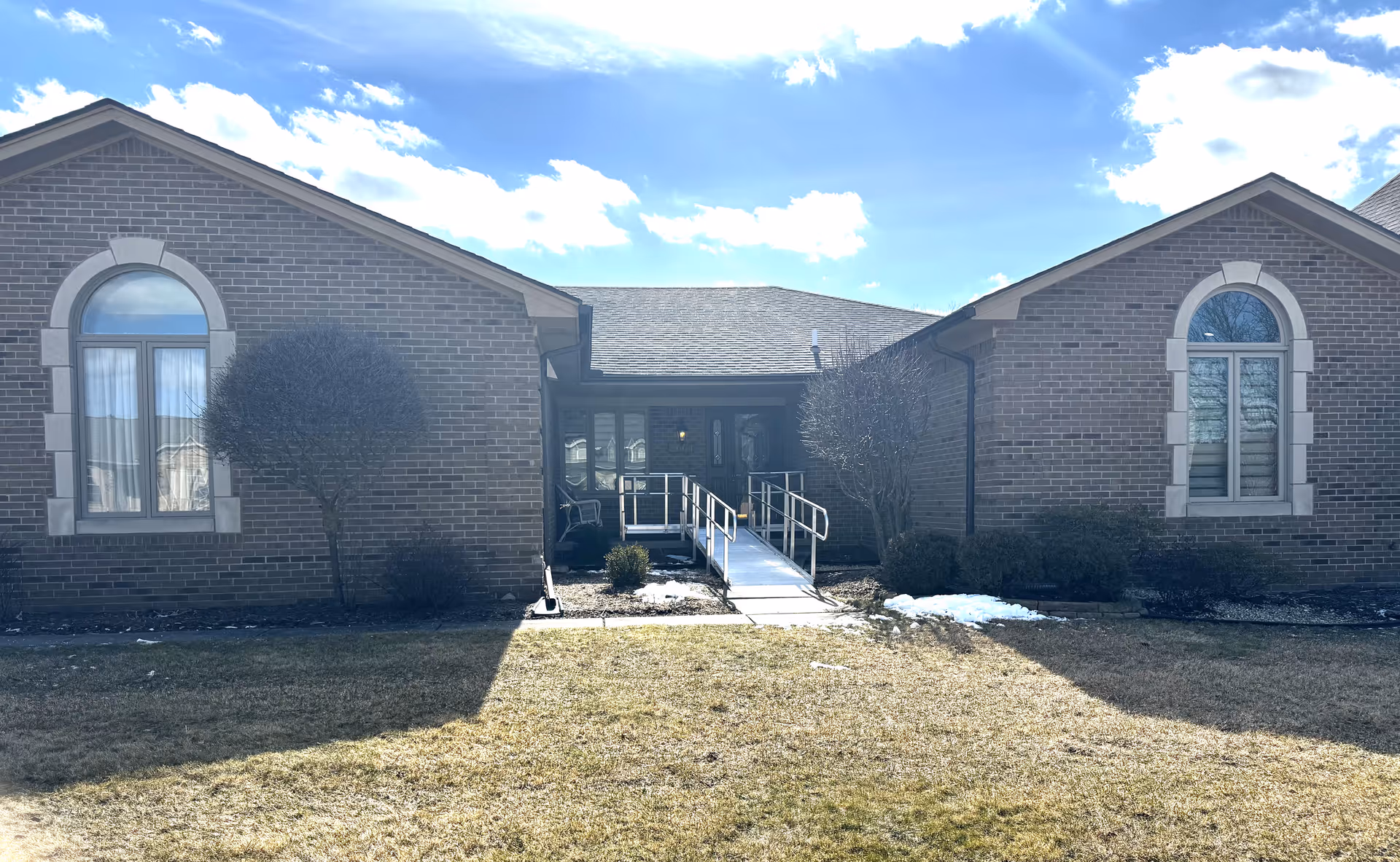 Front exterior of a brick single-story assisted living building with a ramped entrance, arched windows, and a grassy lawn.