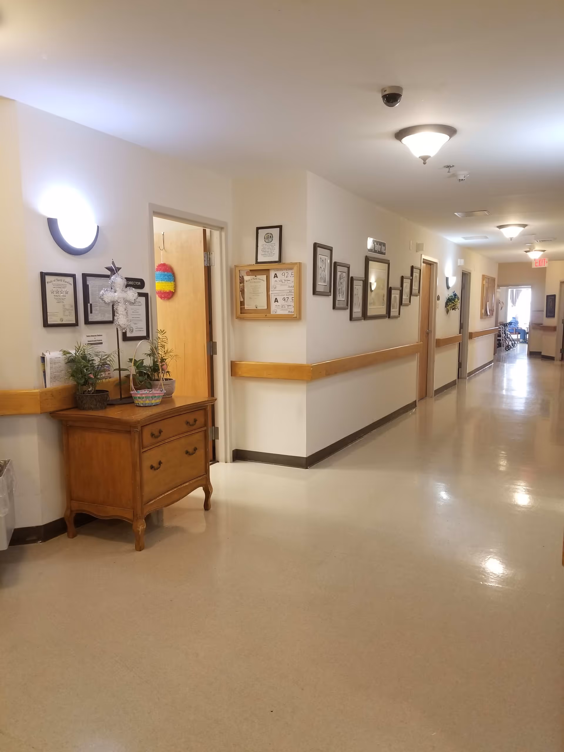 A clean, well-lit hallway in an assisted living facility with beige walls and floor. On the left side, there is a wooden cabinet with plants and a decorative cross on top. Several framed certificates and pictures hang on the walls. The hallway extends into the distance with doors on both sides and an exit sign visible at the far end.