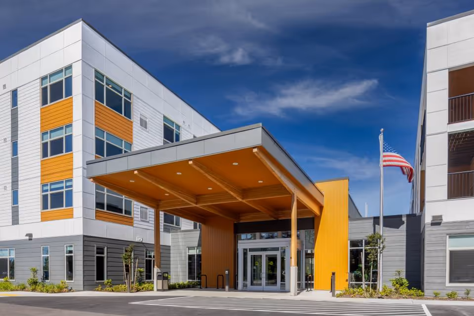 Entrance of a modern multi-story senior living building with a covered canopy, yellow accents, and an American flag.