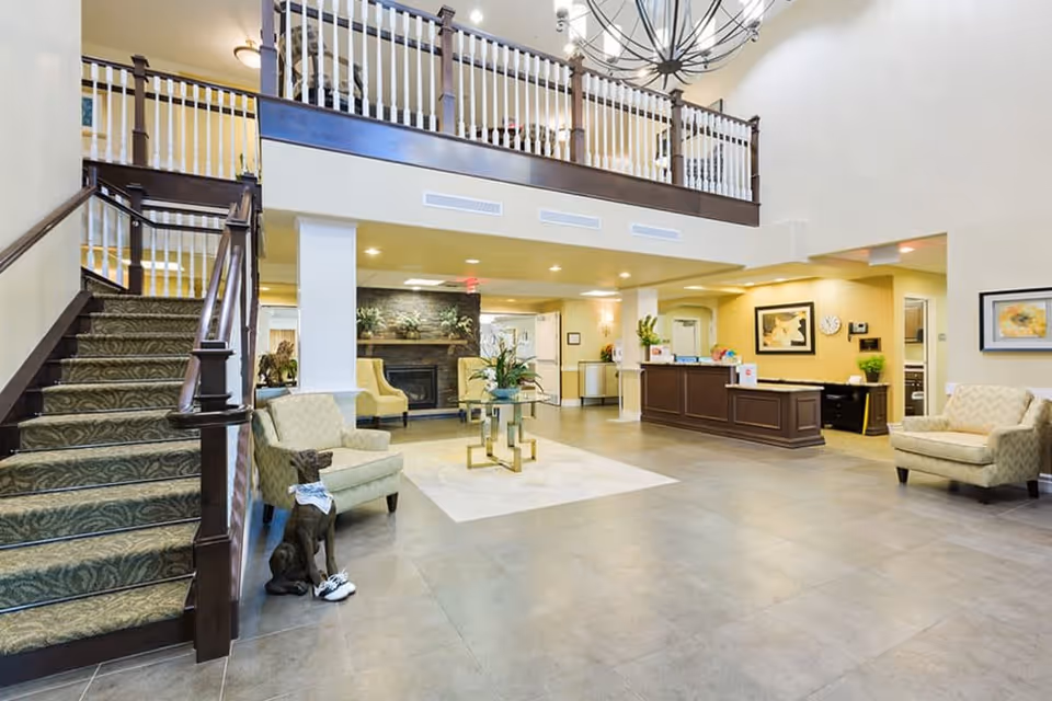 Spacious senior living facility lobby with a staircase on the left, two beige armchairs, a glass coffee table with a plant in the center, a fireplace in the background, and a reception desk on the right. The area is well-lit with ceiling lights and decorated with framed artwork and plants.