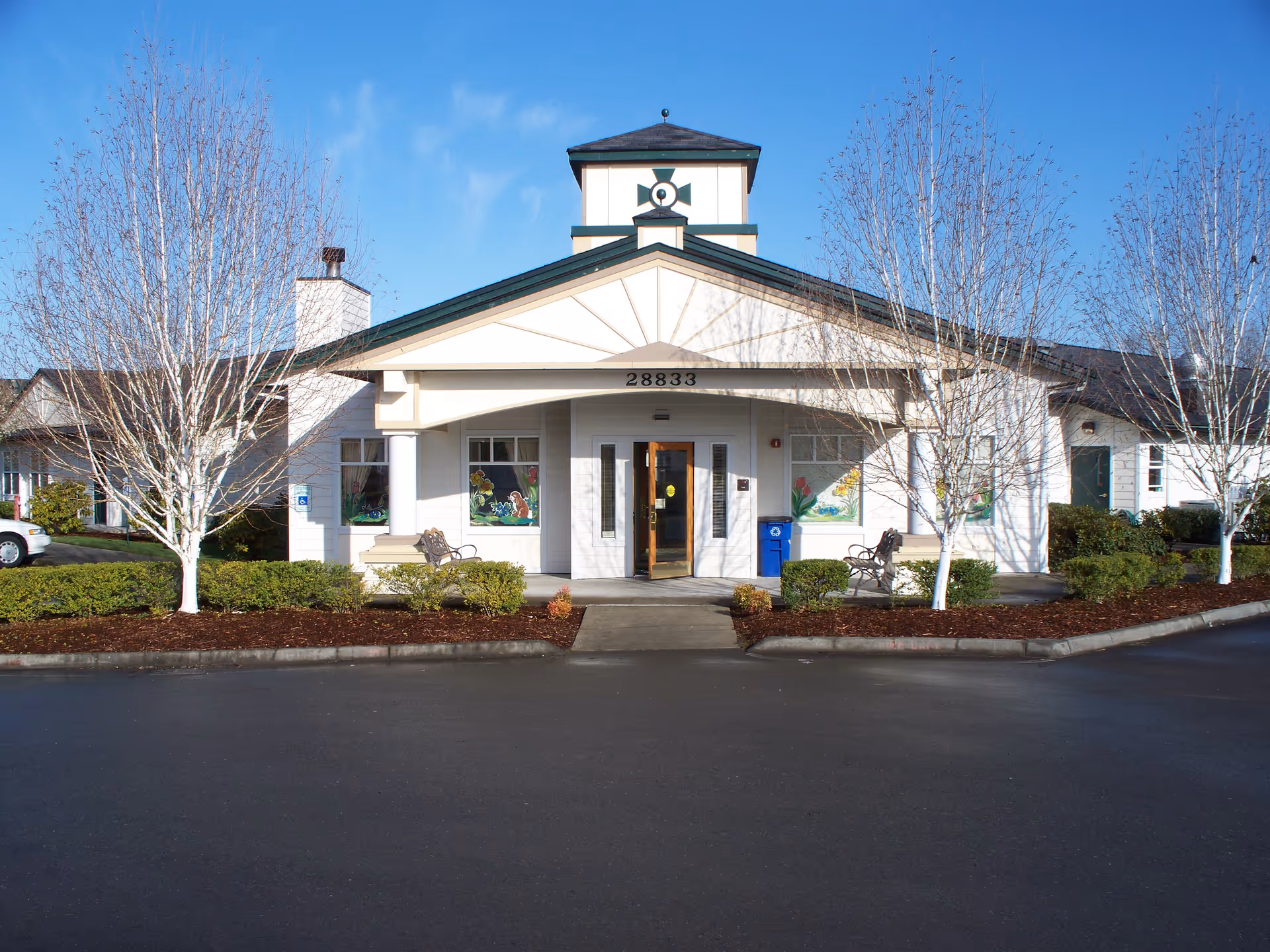 Front exterior view of a single-story assisted living facility building with a peaked roof and a small tower structure on top. The entrance has a covered porch with two benches on either side and decorative windows with colorful artwork. There are leafless trees and shrubs in front of the building, and a clear blue sky above.