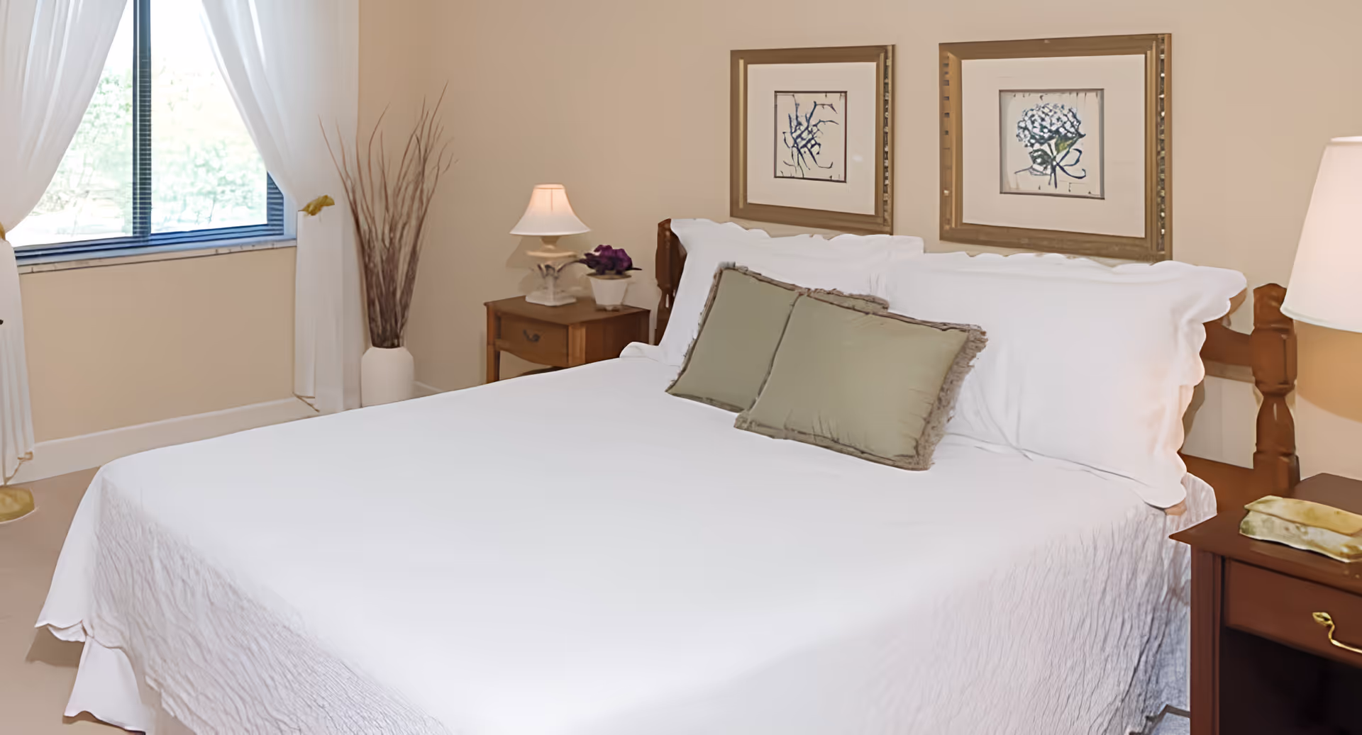 A neatly made bed with white bedding and two green pillows in a bedroom. There are two framed floral artworks hanging on the beige wall above the bed. On each side of the bed are wooden nightstands with lamps and decorative items. A window with white curtains and a vase with tall dried branches are visible on the left side.