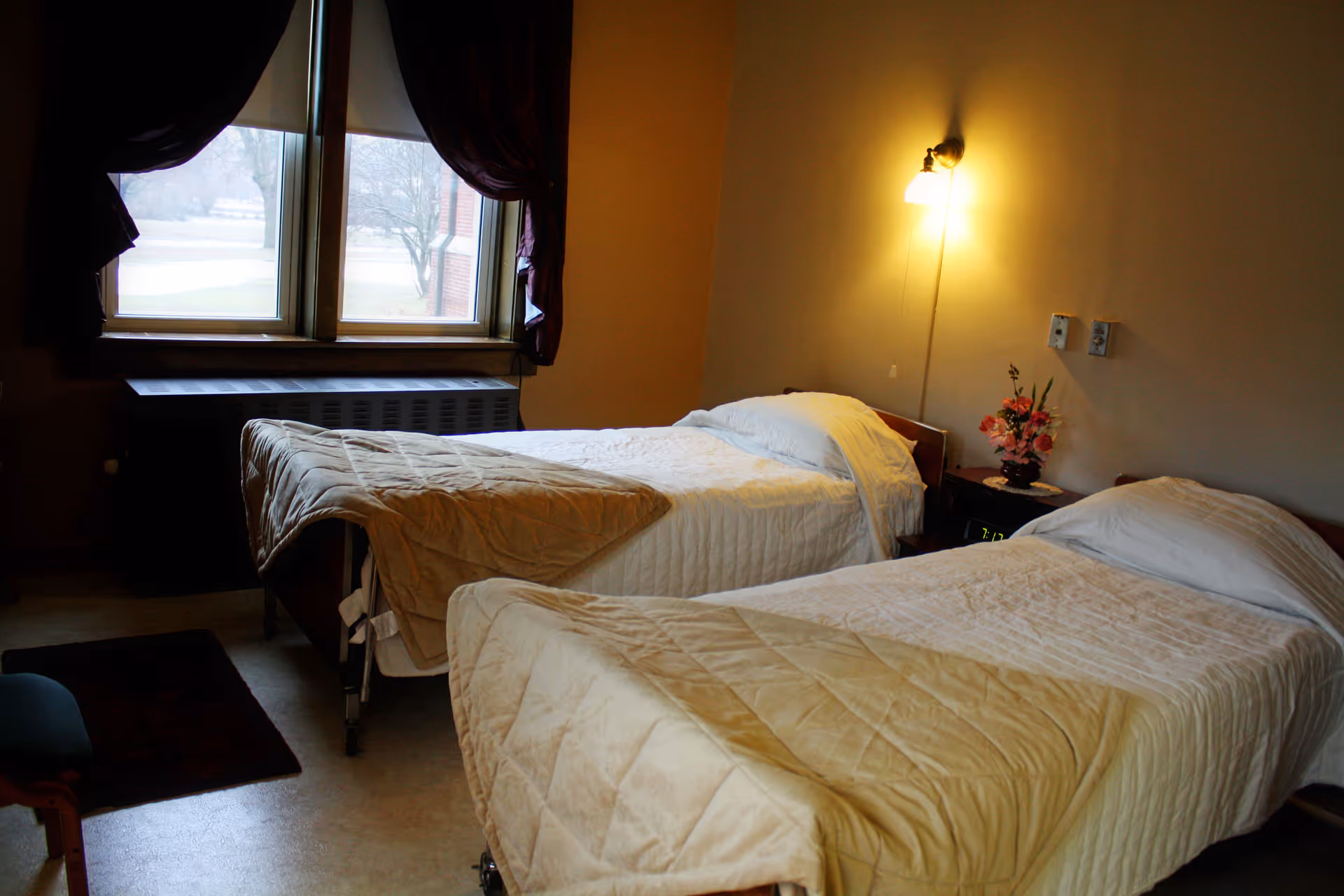 A bedroom with two single beds, each made with white sheets and beige quilts. There is a nightstand between the beds with a small flower arrangement and a clock. A wall-mounted lamp provides warm lighting above the nightstand. A window with dark curtains is visible on the left side of the room.