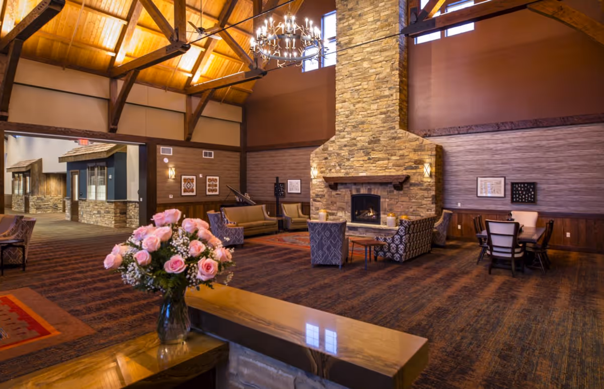 Spacious senior living lounge with a high wooden-beamed ceiling, stone fireplace, seating areas, and a vase of pink roses on a table.