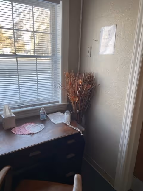 A corner of a room with a window covered by white blinds letting in natural light. Below the window is a dark wooden dresser with a box of tissues, a bottle of hand sanitizer, a salt shaker, and some heart-shaped doilies on top. To the right of the dresser is a tall vase filled with dried decorative plants. A chair with a wooden back is partially visible in the foreground. A small sign is taped to the wall above the vase.