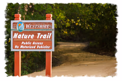 A wooden signpost at the entrance of a nature trail surrounded by green trees. The sign reads 'Westshire Nature Trail Public Access No Motorized Vehicles'. A dirt path leads into the wooded area.