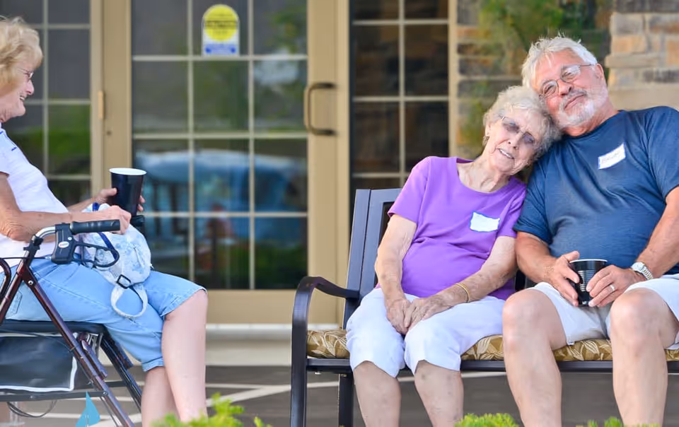 Three elderly individuals sitting outside a building entrance. One woman is seated on a walker with a cup in her hand, while another woman and a man sit closely together on a bench, smiling and holding cups. The background shows glass doors and a stone wall.