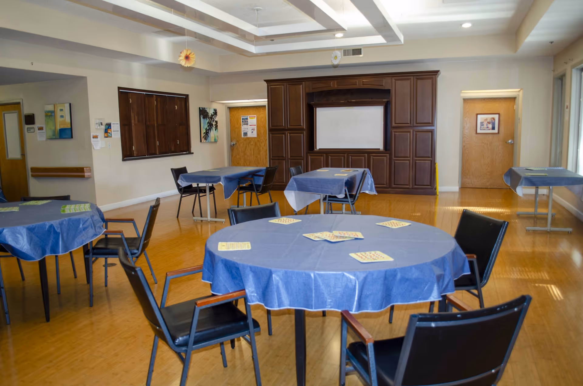 A community room with several tables covered in blue tablecloths and black chairs arranged around them. The tables have bingo cards placed on top. The room has wooden flooring, beige walls, and a large wooden cabinet with a whiteboard in the center. There are two wooden doors and some wall decorations including paintings and hanging paper flowers.