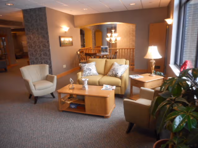 Cozy common living area with a sofa, two armchairs, coffee table and side table by a window, with a dining area visible in the background.