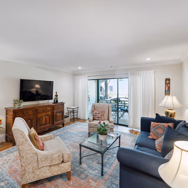 A cozy living room with a blue sofa, two beige armchairs, a glass coffee table with a plant on it, and a wooden cabinet with a flat-screen TV mounted above it. The room has a large window with white curtains leading to an outdoor patio with a table and chairs. The floor is covered with a patterned area rug, and there is a table lamp on a side table next to the sofa.