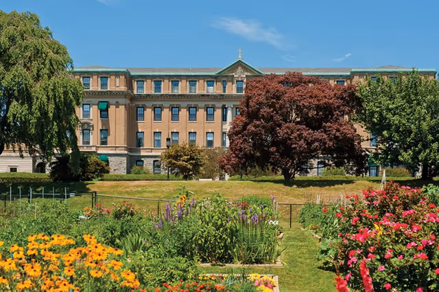 A large, multi-story brick building with a cross on top, viewed from a garden filled with colorful flowers and greenery under a clear blue sky.