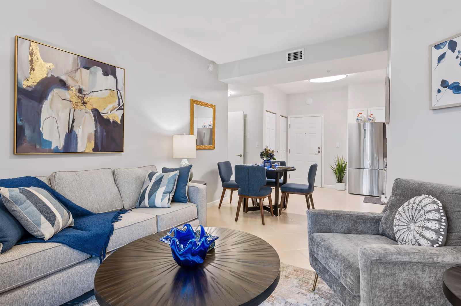 A modern living room and dining area with a light gray sofa adorned with blue and striped pillows, a gray armchair with a decorative round pillow, and a dark round coffee table with a blue glass centerpiece. The dining area features a round table with four blue upholstered chairs. The walls are light gray with abstract artwork and a mirror. A stainless steel refrigerator is visible in the background near the entrance door.