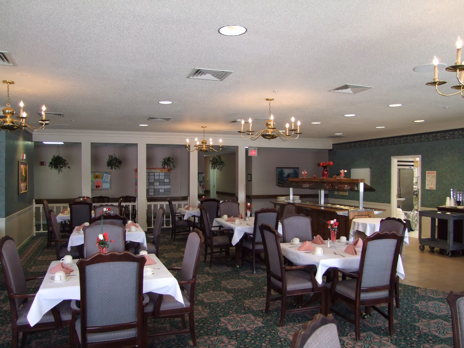 Dining room with multiple tables covered with white tablecloths, each set with cups, napkins, and small flower arrangements. The room has green patterned carpet, green wallpaper, and brass chandeliers hanging from the ceiling. There is a buffet counter on the right side and a doorway leading to another room.