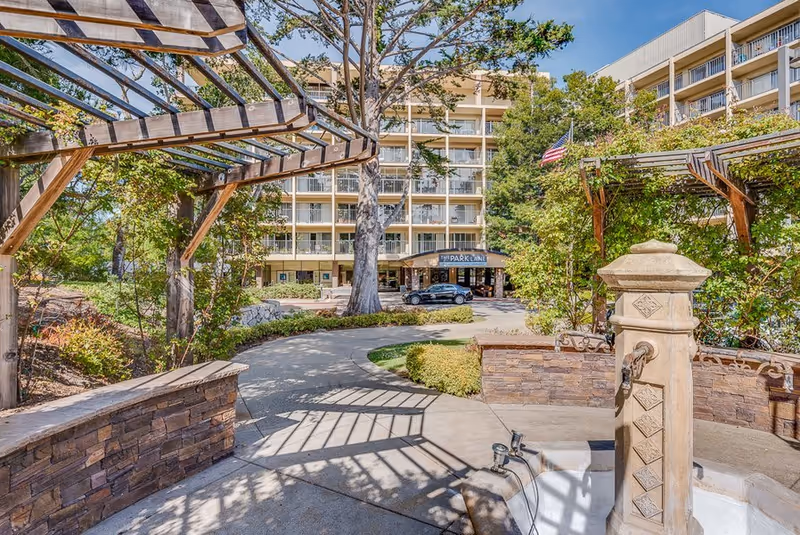 Outdoor garden area with a stone water fountain and wooden pergolas casting shadows on the pathway, leading to the entrance of a multi-story senior living facility named The Park Lane, surrounded by trees and greenery under a clear blue sky.
