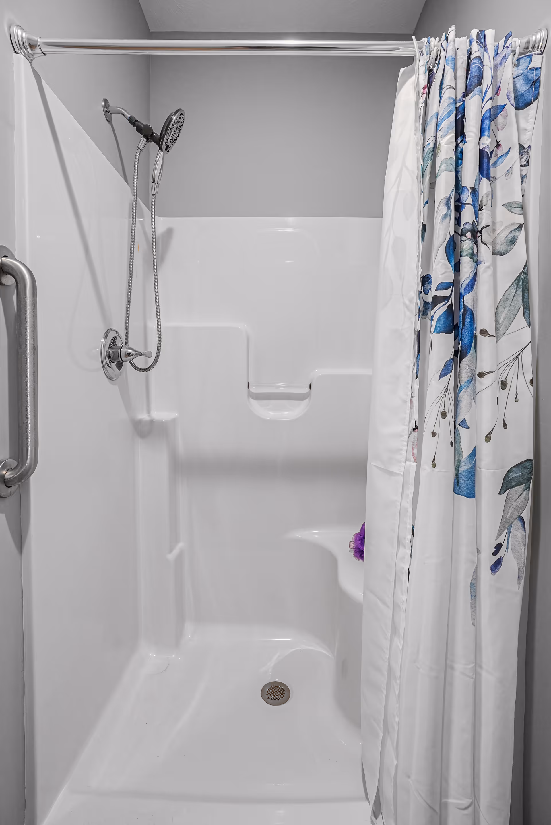 A clean white shower stall with a built-in seat and a handheld showerhead mounted on the wall. There is a silver grab bar on the left side and a shower curtain with a blue floral pattern partially drawn to the right.