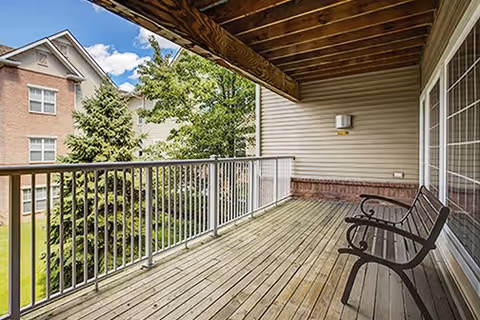 A wooden balcony with two black metal chairs, overlooking a grassy area with trees and a multi-story brick building in the background under a partly cloudy sky.