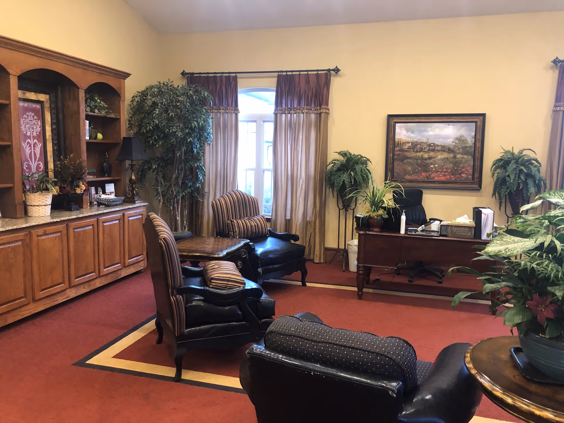 A cozy office or lounge area with a red carpet and beige walls. The room features a wooden desk with office supplies, two large potted plants on either side of the desk, and a framed landscape painting on the wall behind it. There are three upholstered chairs with striped cushions arranged around a wooden coffee table. A large wooden cabinet with shelves and decorative items is on the left side of the room, and a window with beige curtains is in the center background.