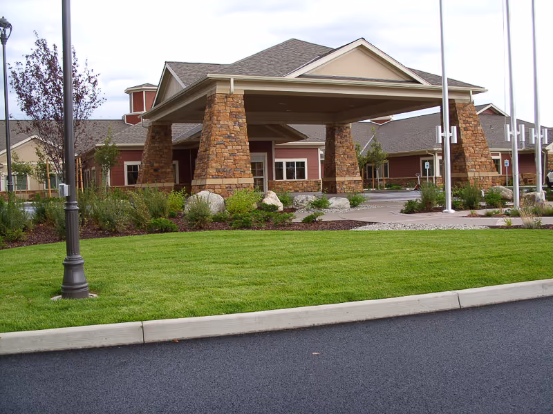 Entrance of a senior living facility with a covered drop-off area supported by stone pillars, surrounded by landscaped greenery and a well-maintained lawn, with a paved driveway in front.