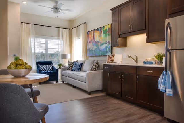 A cozy living area with a gray sofa adorned with patterned cushions, a blue armchair, a round wooden dining table with chairs, and a kitchenette with dark wood cabinets and a stainless steel refrigerator. The room is well-lit with natural light coming through large windows covered with white curtains, and a colorful abstract painting hangs on the wall above the sofa.