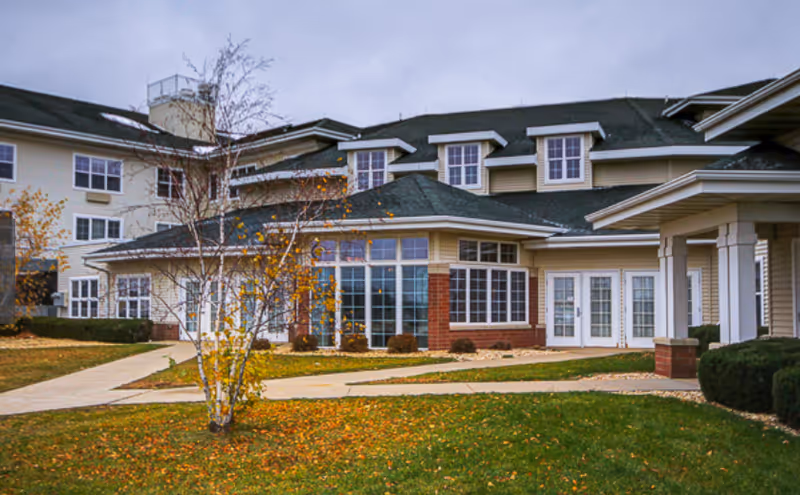 Exterior view of a multi-story senior living facility building with beige siding, multiple windows, and a green roof. There is a paved walkway leading to the entrance, surrounded by grass and a few small trees with autumn leaves.