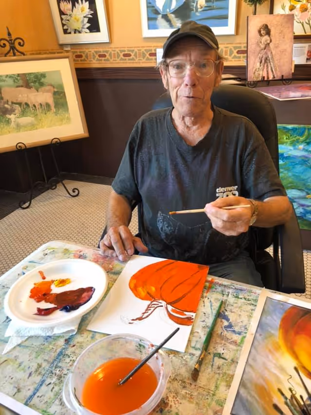 An elderly man wearing glasses and a cap is seated at a table painting an orange pumpkin on a piece of paper. The table is covered with paint supplies including a plate with various paint colors and a bowl of orange water with a brush in it. Behind him are several framed paintings displayed on the wall and on stands.