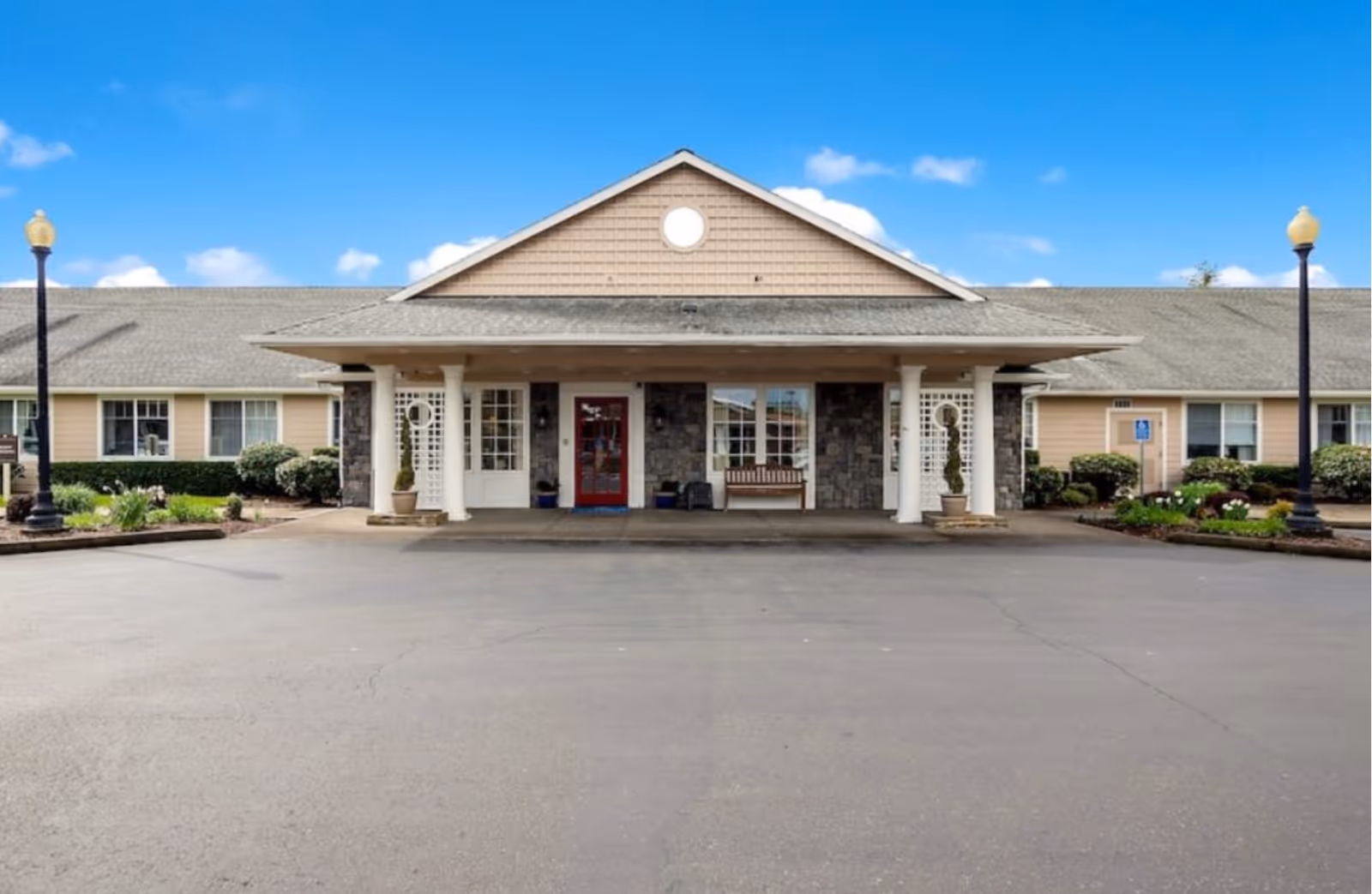 Front exterior view of McMinnville Memory Care facility showing a single-story building with a covered entrance supported by white columns, a red door, windows, two lamp posts, and landscaped greenery under a clear blue sky.