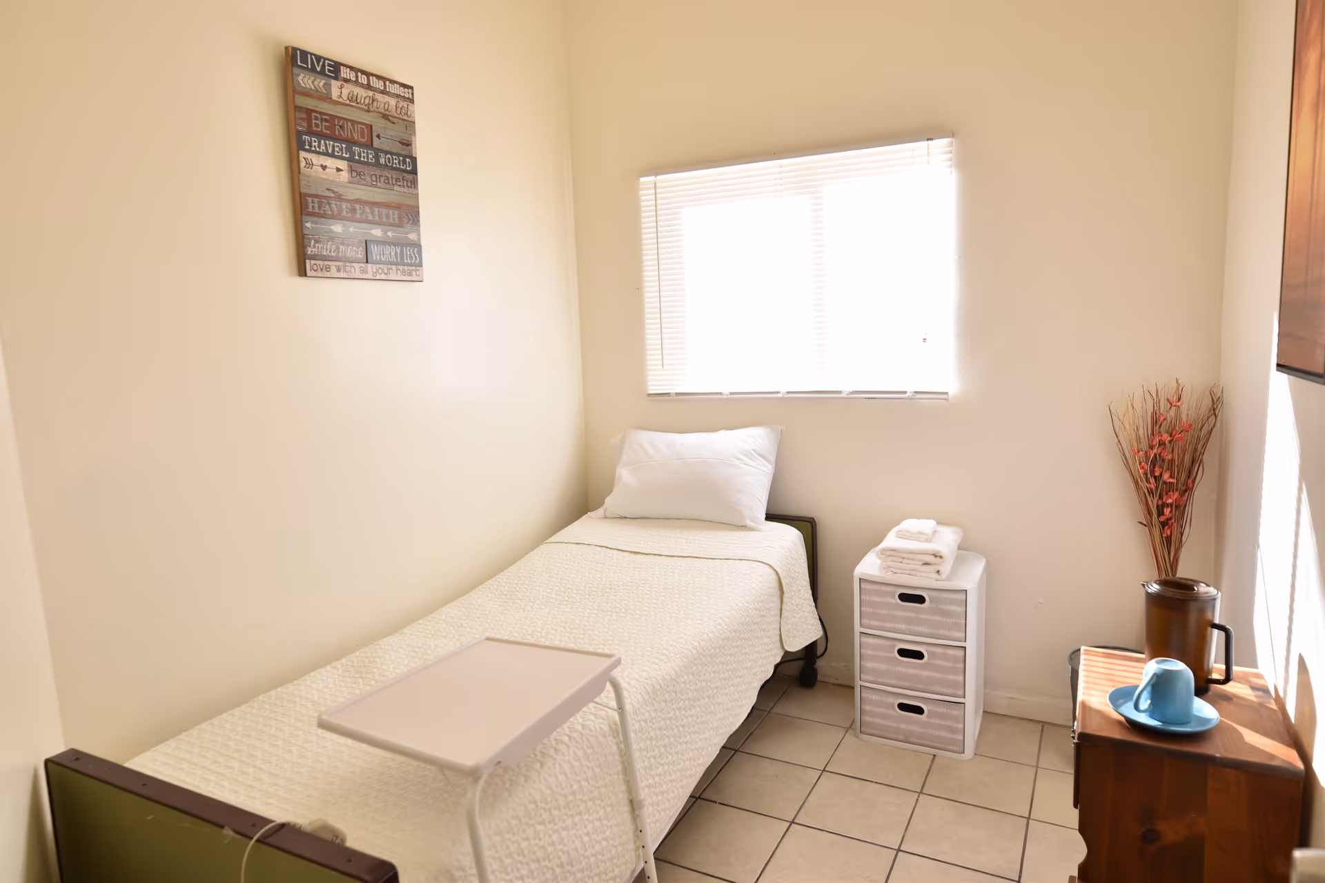 Small bright bedroom with a single bed, a window with blinds, a three-drawer storage unit, and a nightstand with a cup.
